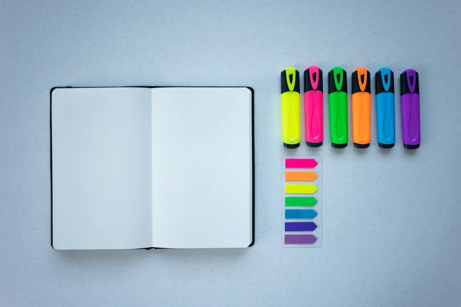 An open notebook next to a row of colored highlighter pens on a desk